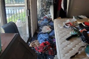 Bedroom left with a bare mattress, clothes and trash across the floor and a shattered glass door